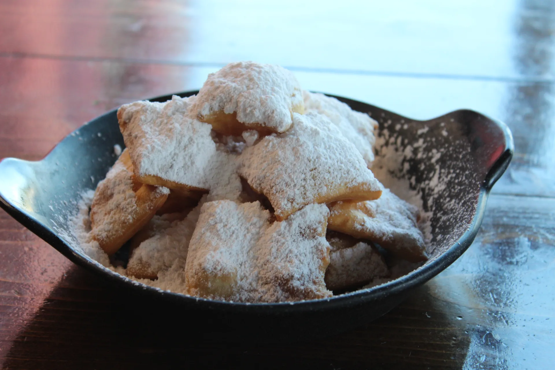 Golden beignet bites dusted with powdered sugar in a black bowl, ready to be enjoyed.
