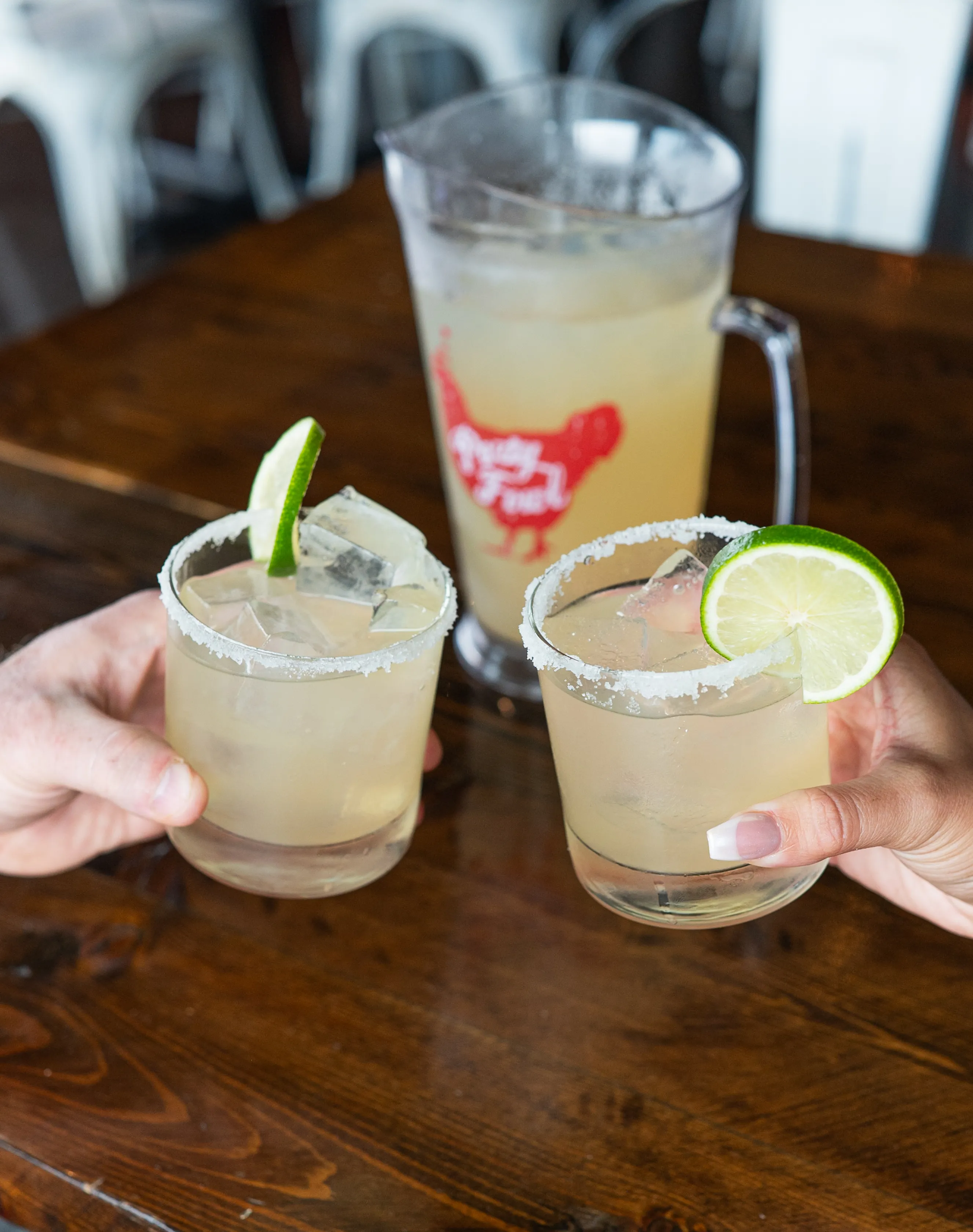 Two hands holding margaritas with lime slices and salted rims, with a pitcher of margarita in the background.