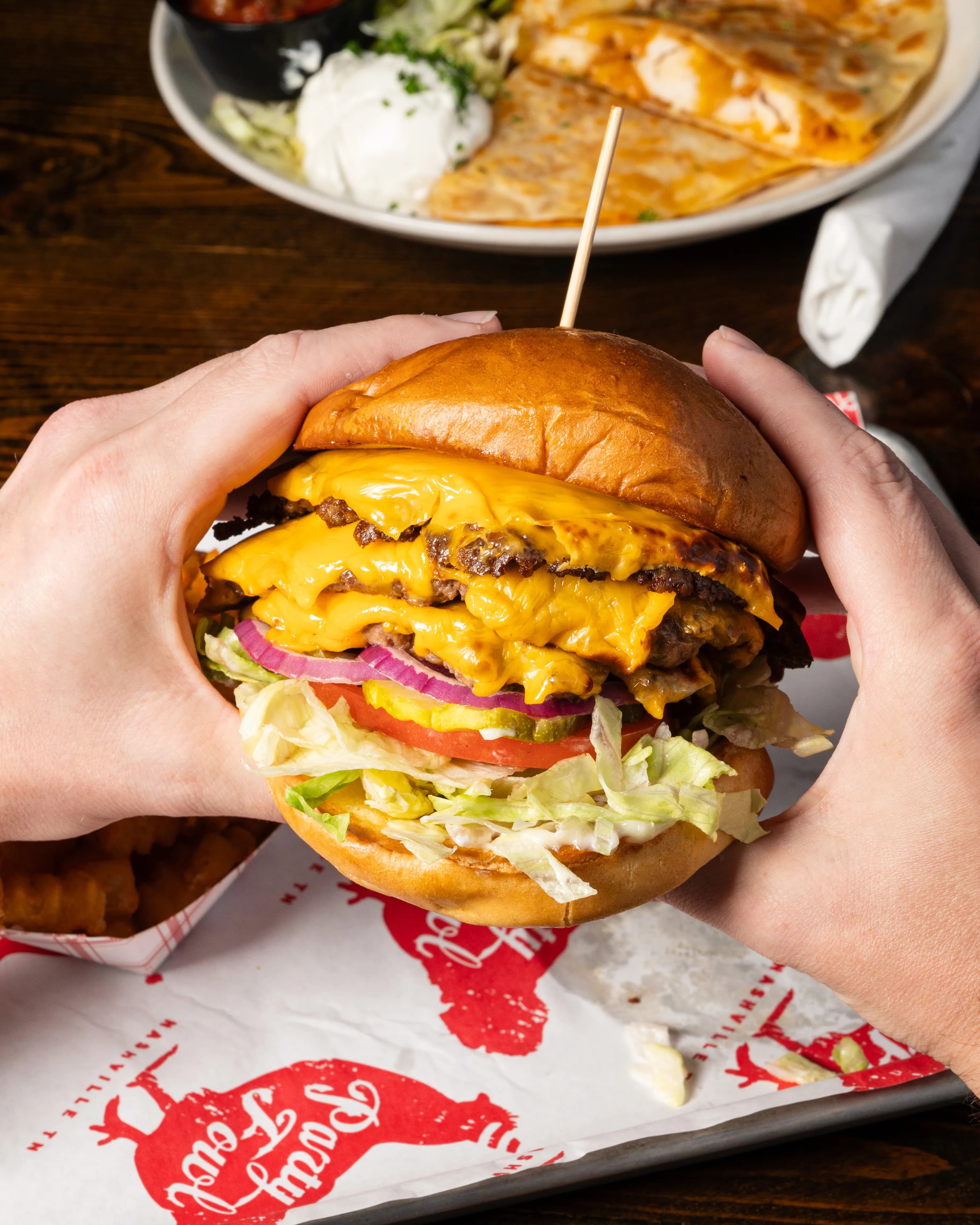 A hand holding a towering triple cheeseburger with layers of beef, melted cheese, lettuce, tomato, and red onion.