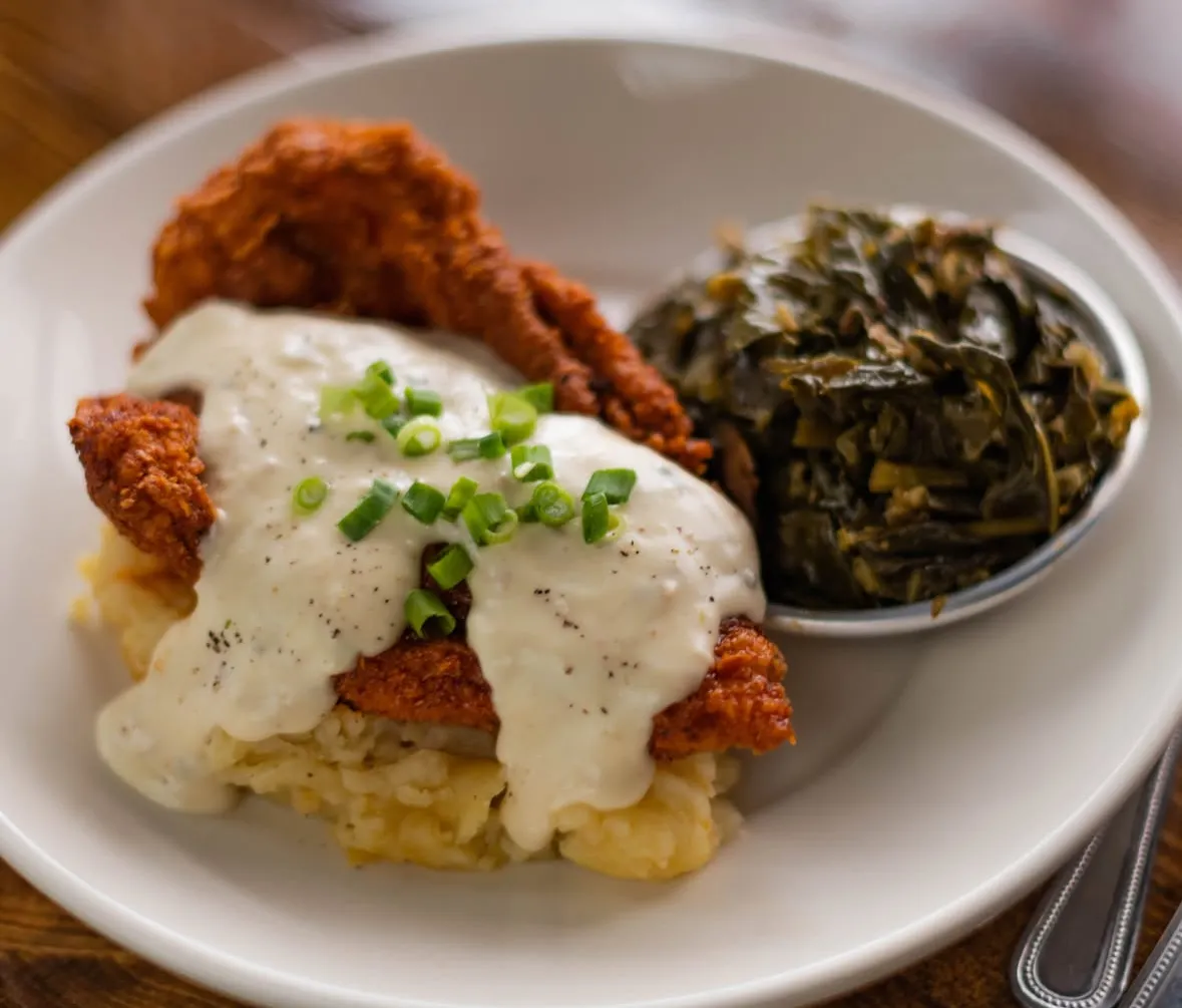 A plate featuring fried chicken topped with creamy gravy, served with mashed potatoes and collard greens.