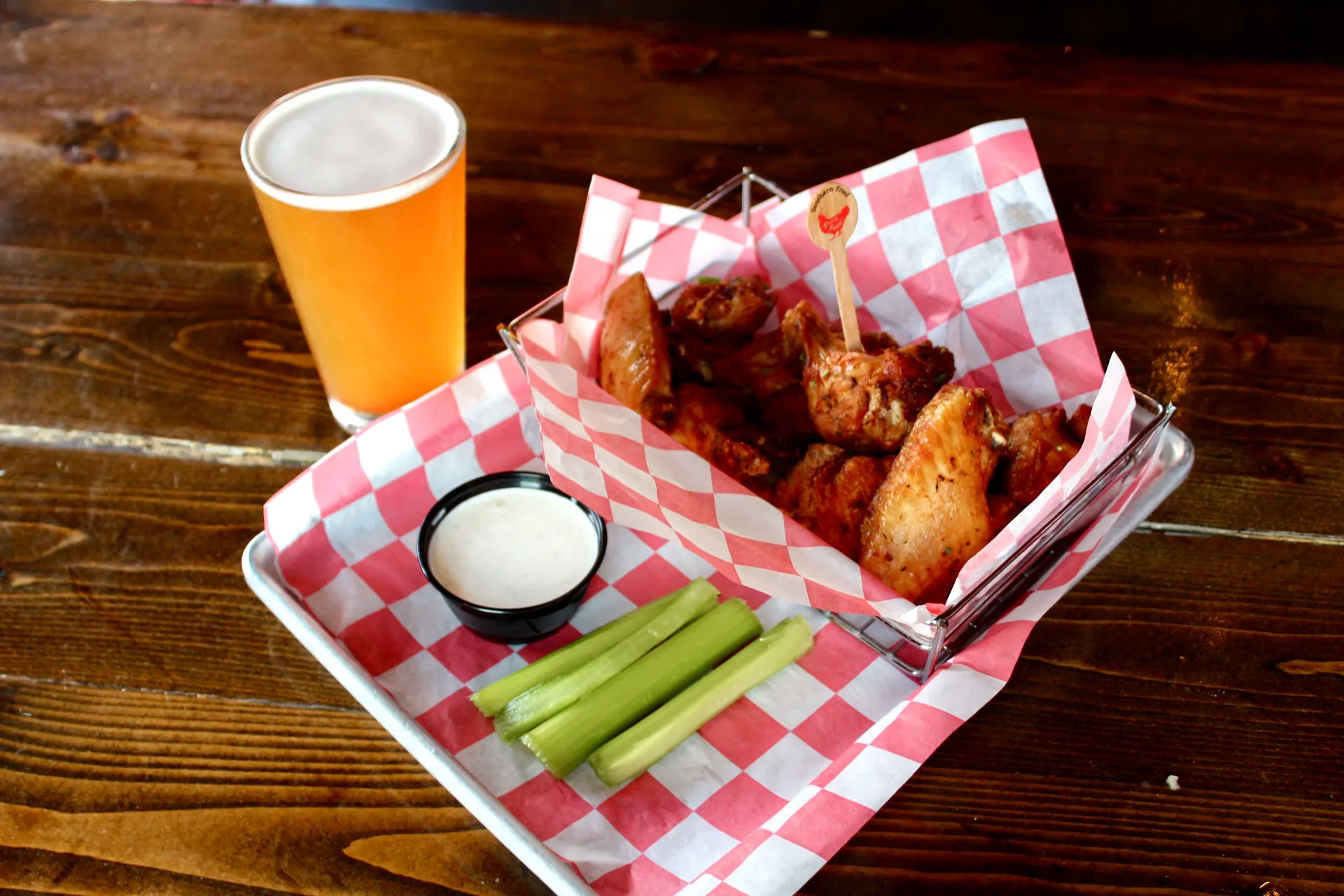 12 Wings A basket of 12 spicy chicken wings served with celery sticks and ranch dressing, accompanied by a glass of beer on a wooden
