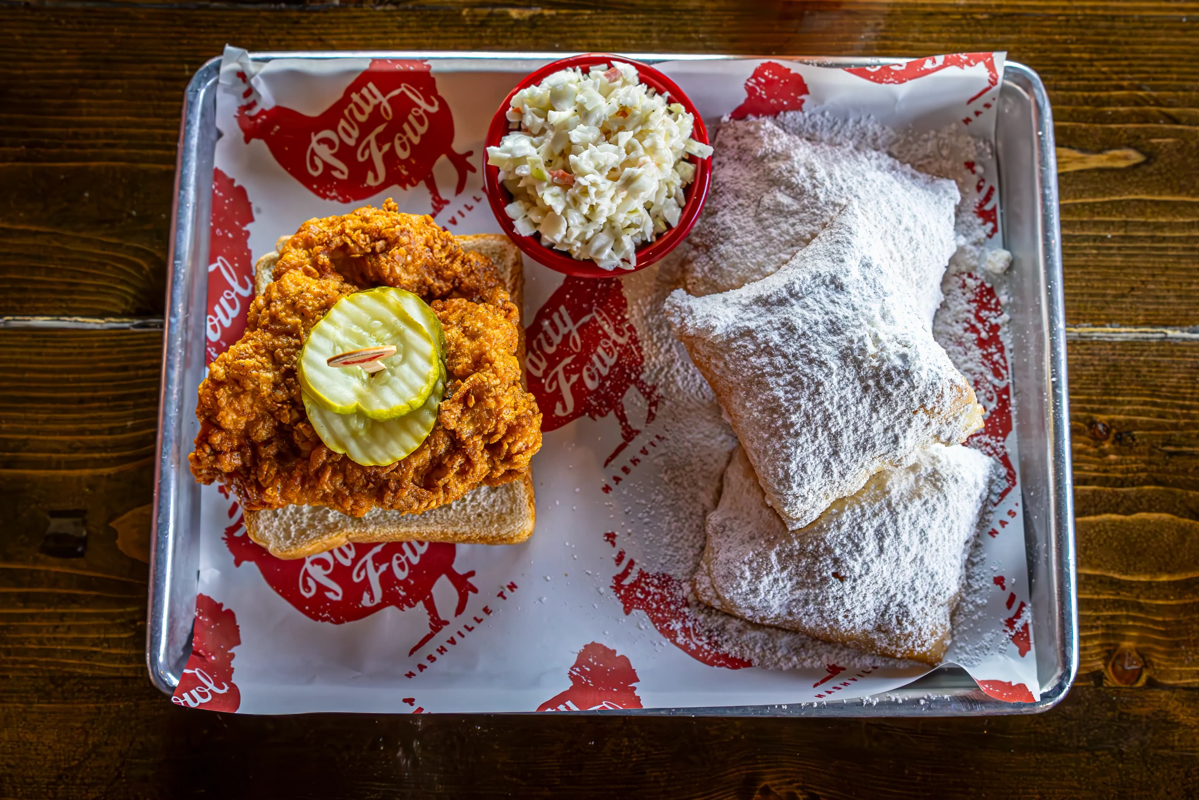 Hot Chicken & Beignets 3 A tray featuring a crispy fried chicken sandwich with pickles, a side of coleslaw, and powdered sugar-covered beignets.