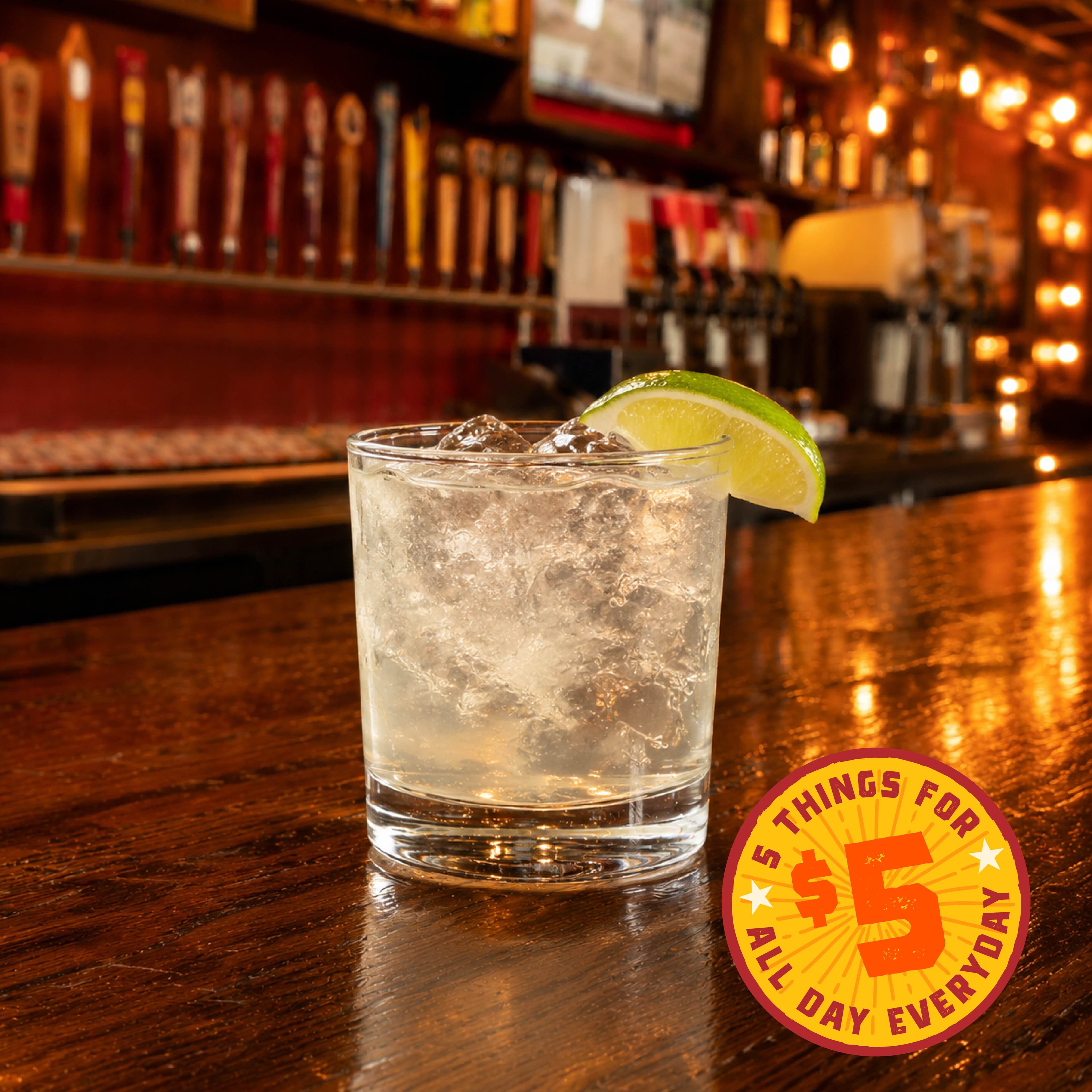 PF Happy Hour Image 01 A refreshing cocktail garnished with a lime slice, served on a bar counter with a promotional sign in the foreground.