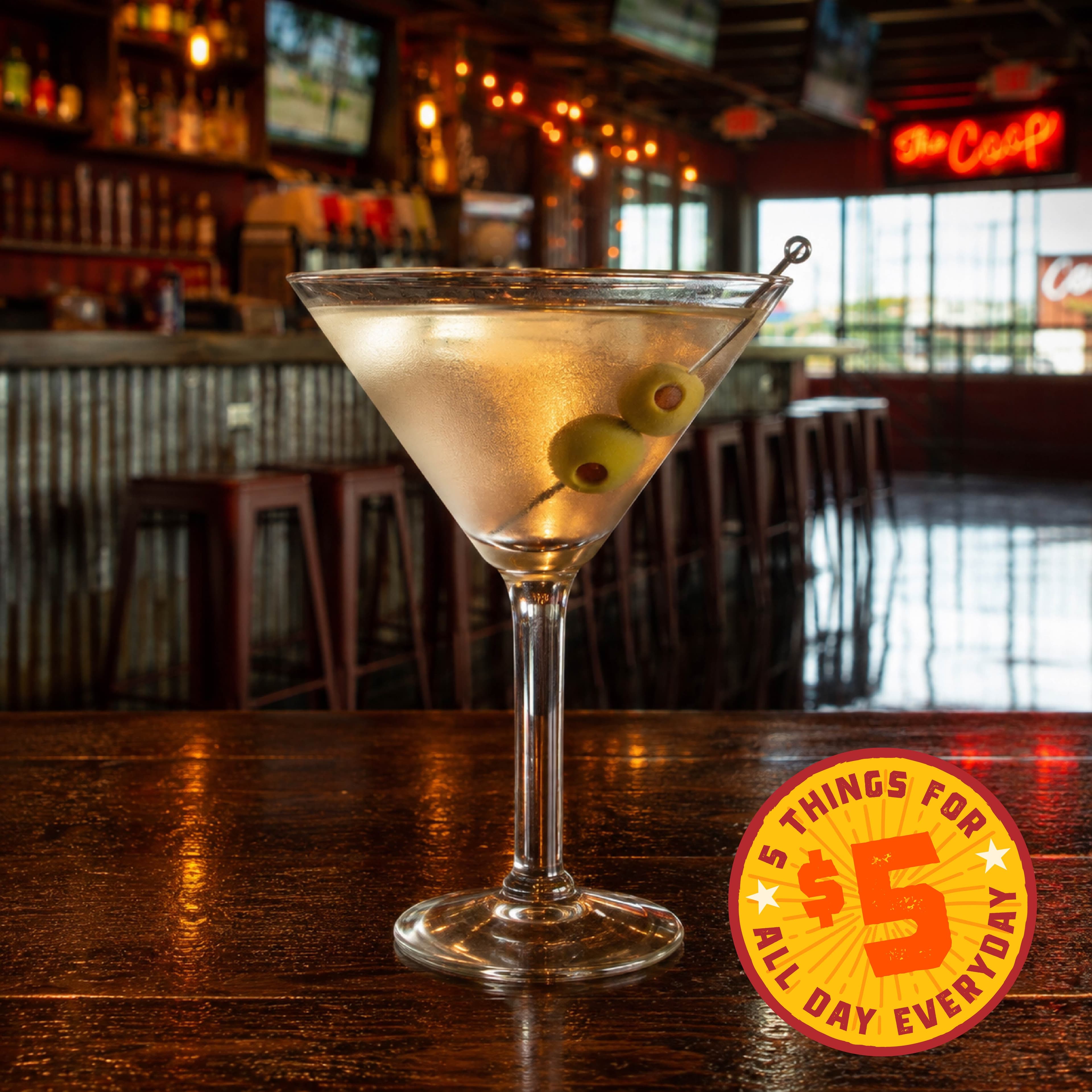 Happy Hour Image 03 A classic martini garnished with olives, served in a dimly lit bar setting with a promotional sign for $5 deals.
