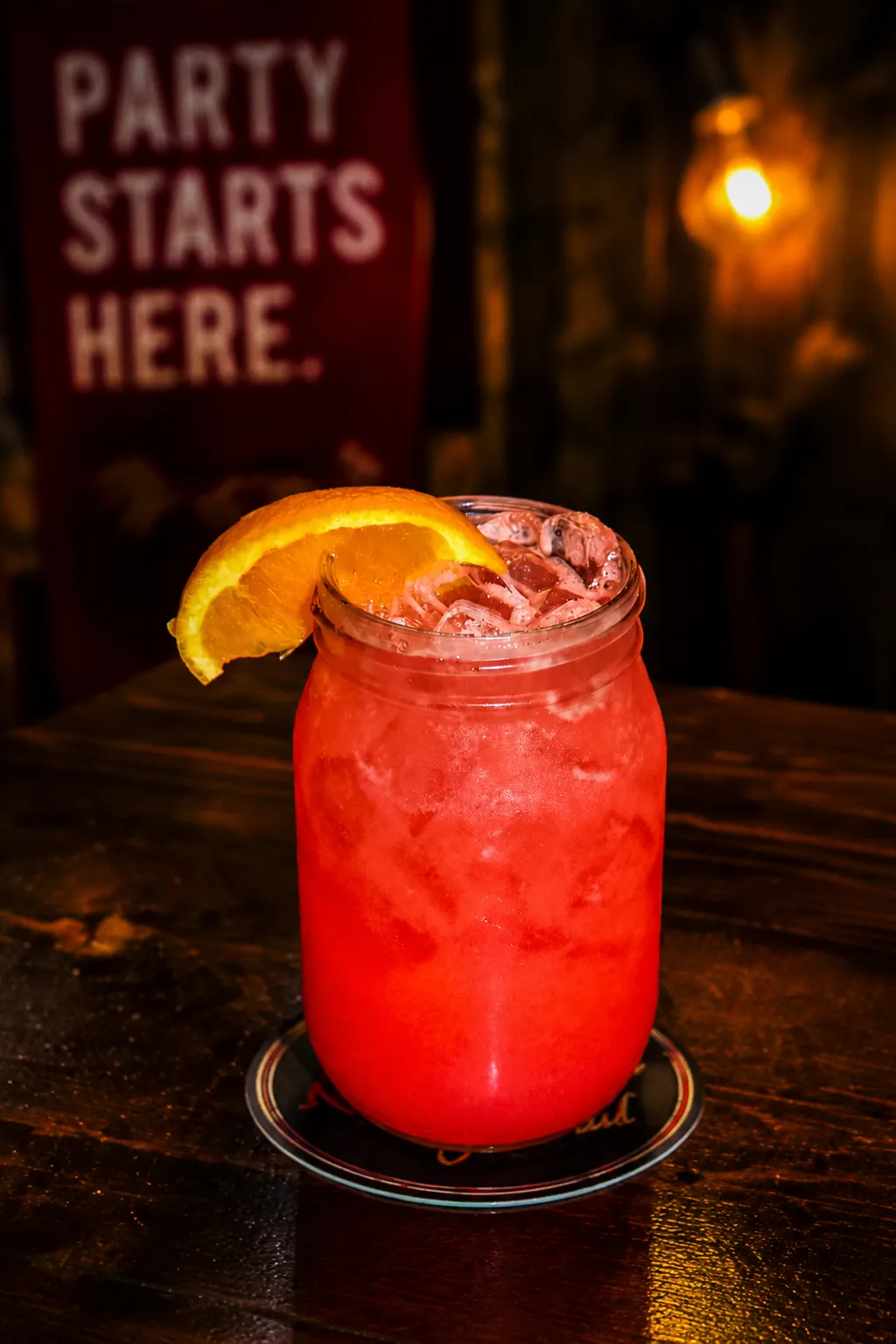 Pain Killer A vibrant red cocktail garnished with an orange slice, served in a mason jar with ice, set against a warm, inviting bar