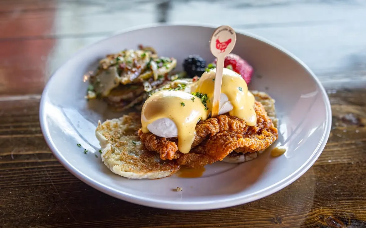 Benedict A plate of crispy fried chicken topped with poached eggs and hollandaise sauce, garnished with herbs and berries.