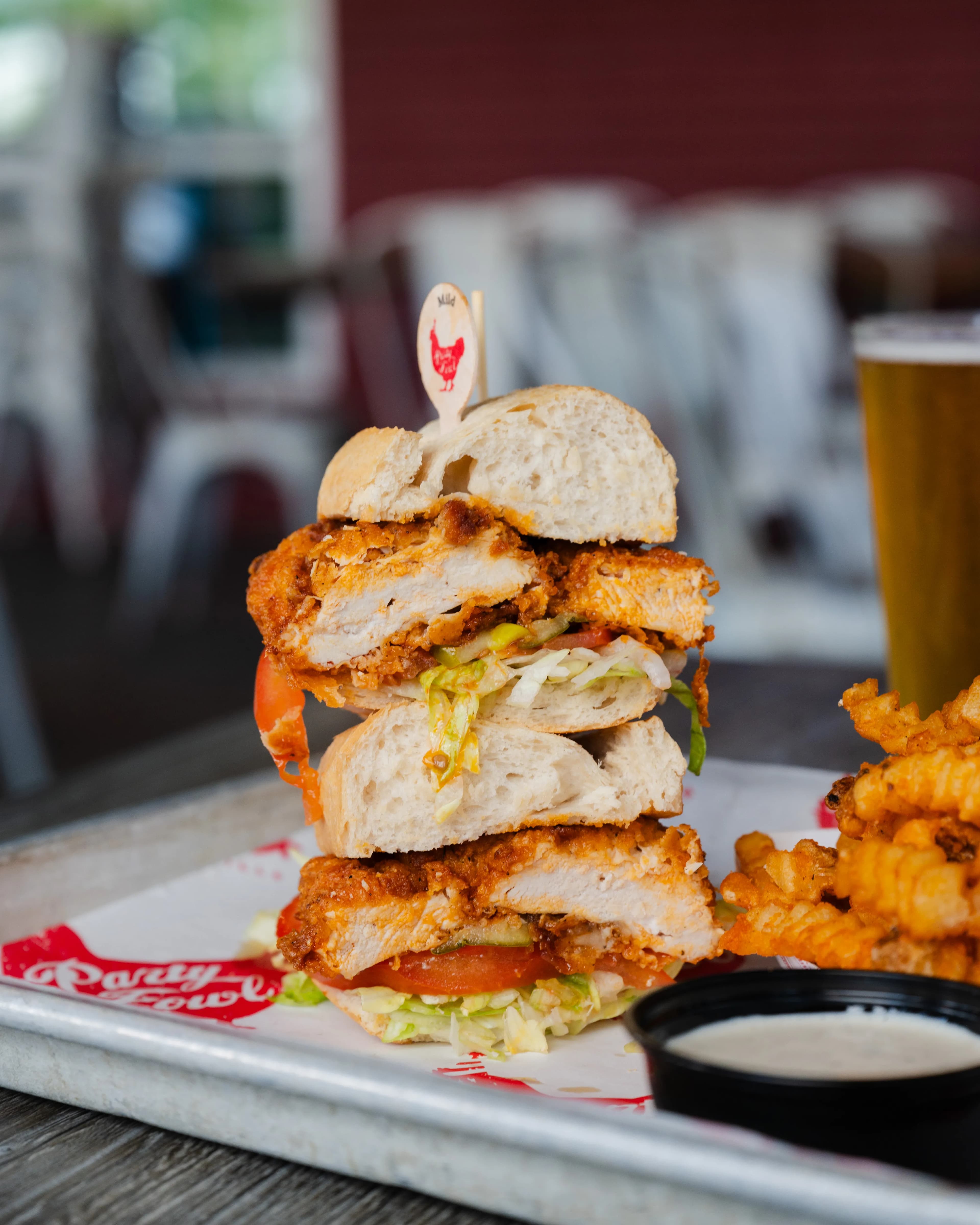 Chicken Po'Boy A towering sandwich with crispy fried chicken, lettuce, tomato, and a side of curly fries on a metal tray.