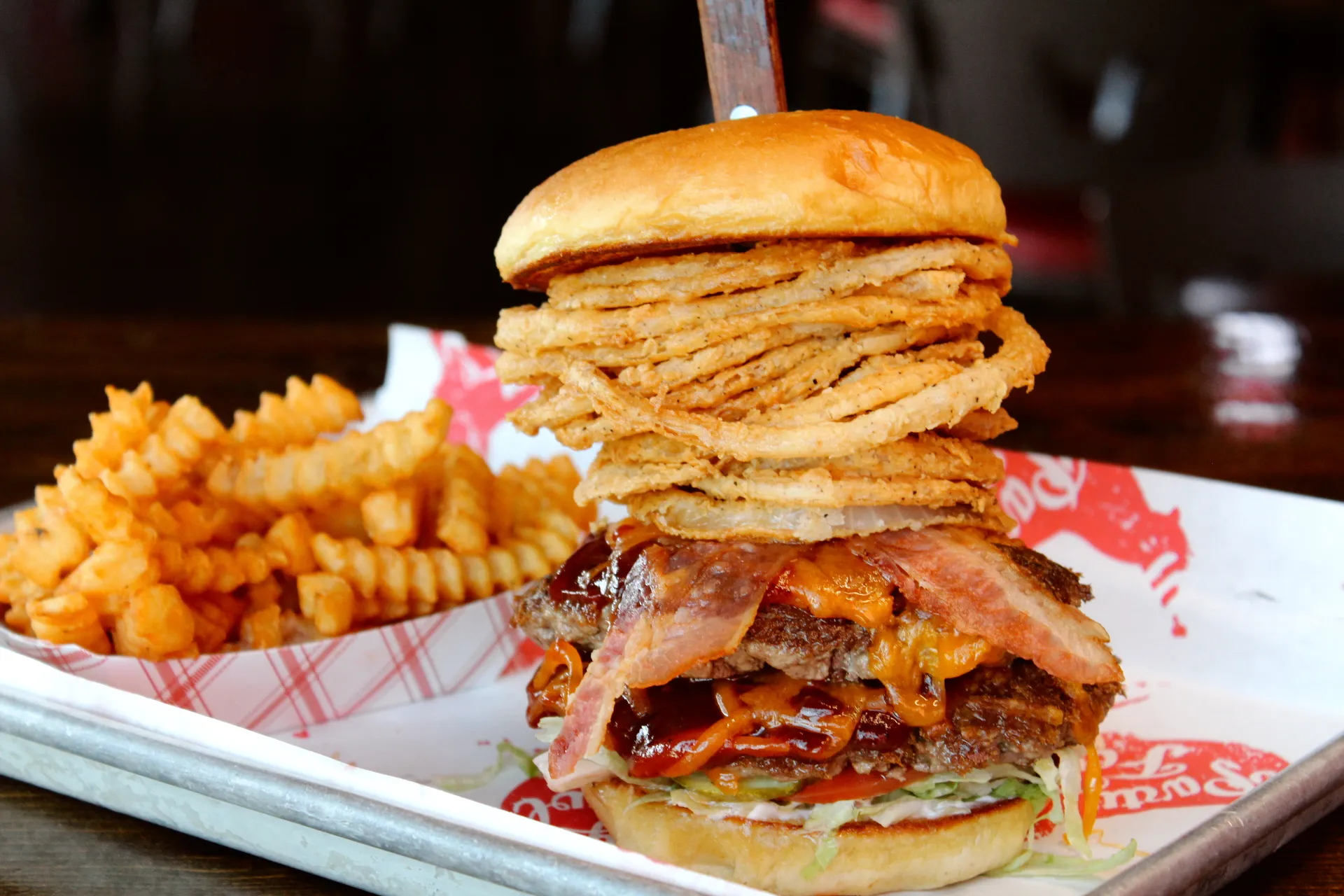 Cowboyburger A towering burger with crispy fried onions, bacon, and cheese, served with curly fries on a metal tray.
