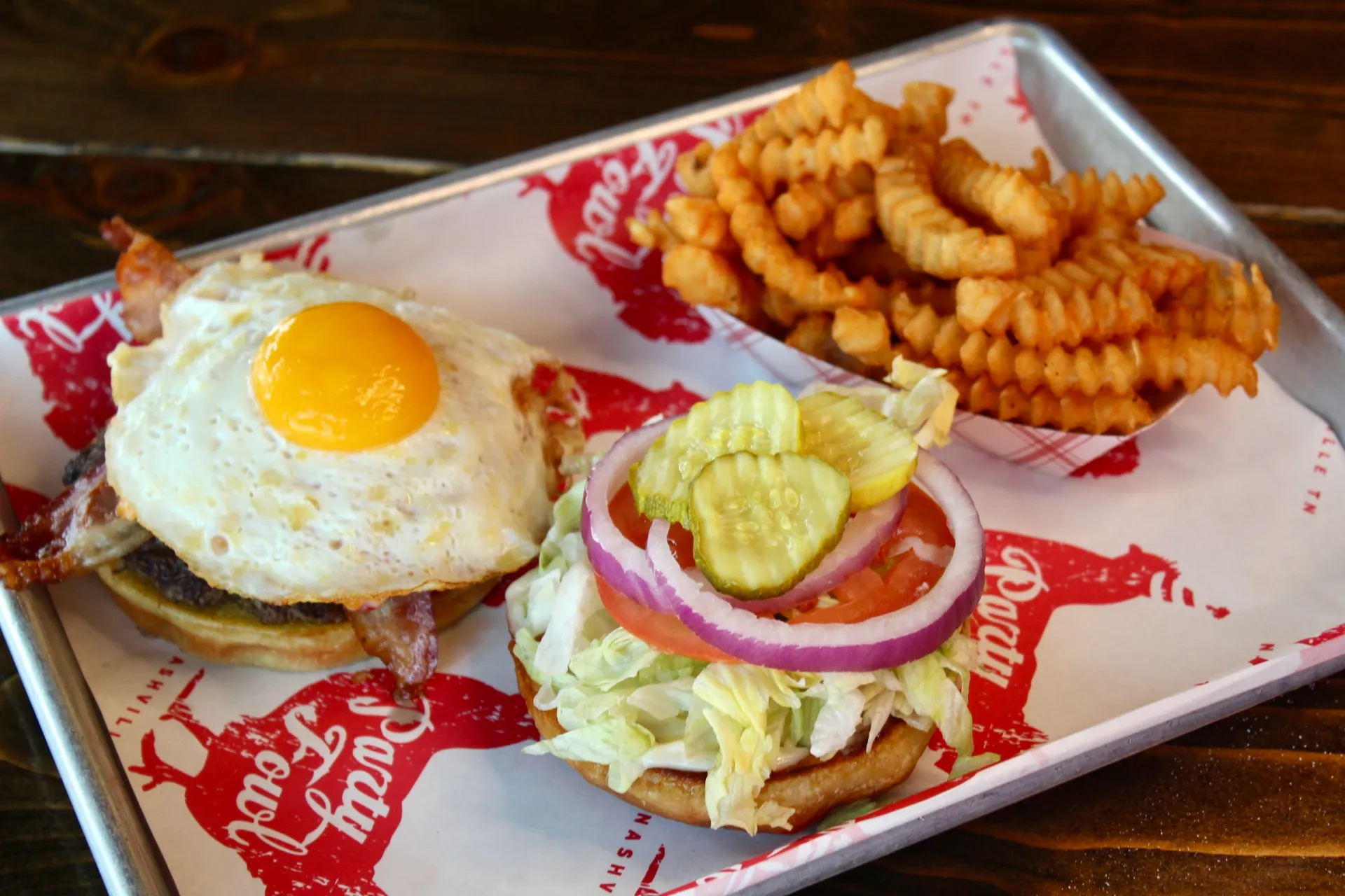 Hangover Burger A hearty meal featuring a burger topped with a sunny-side-up egg, bacon, and fresh toppings, served with crispy waffle fries.