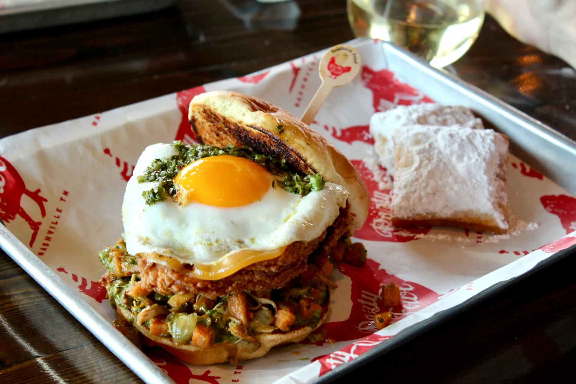 Hen Hash Sandwich A hearty sandwich topped with a sunny-side-up egg and green sauce, served with a side of powdered sugar-dusted donuts.