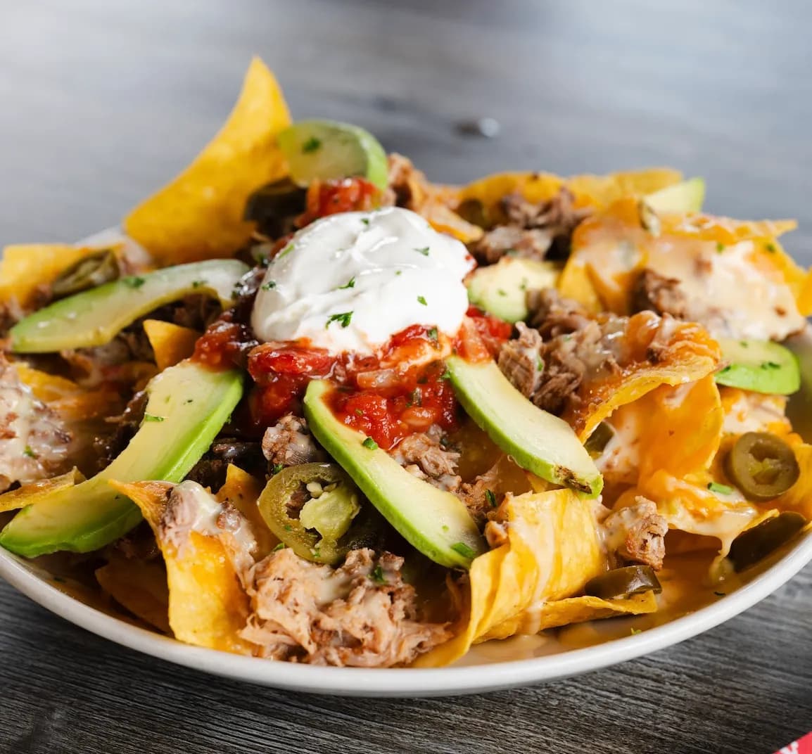 Loadedporknachos A plate of loaded pork nachos topped with avocado slices, sour cream, and salsa, served on a wooden table.